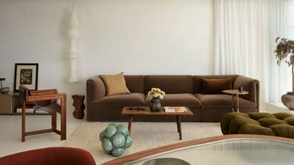 A white living room with a brown couch, a green lounge chair, a white lantern, a wood armchair, a coffee table, and a potted tree