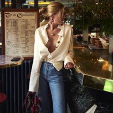 French-British style influencer Camille Charriere poses in a Paris cafe bar wearing red drop earrings, layered necklaces with a yellow-gold pendants, an oversize cream button-down shirt, and high-waisted jeans