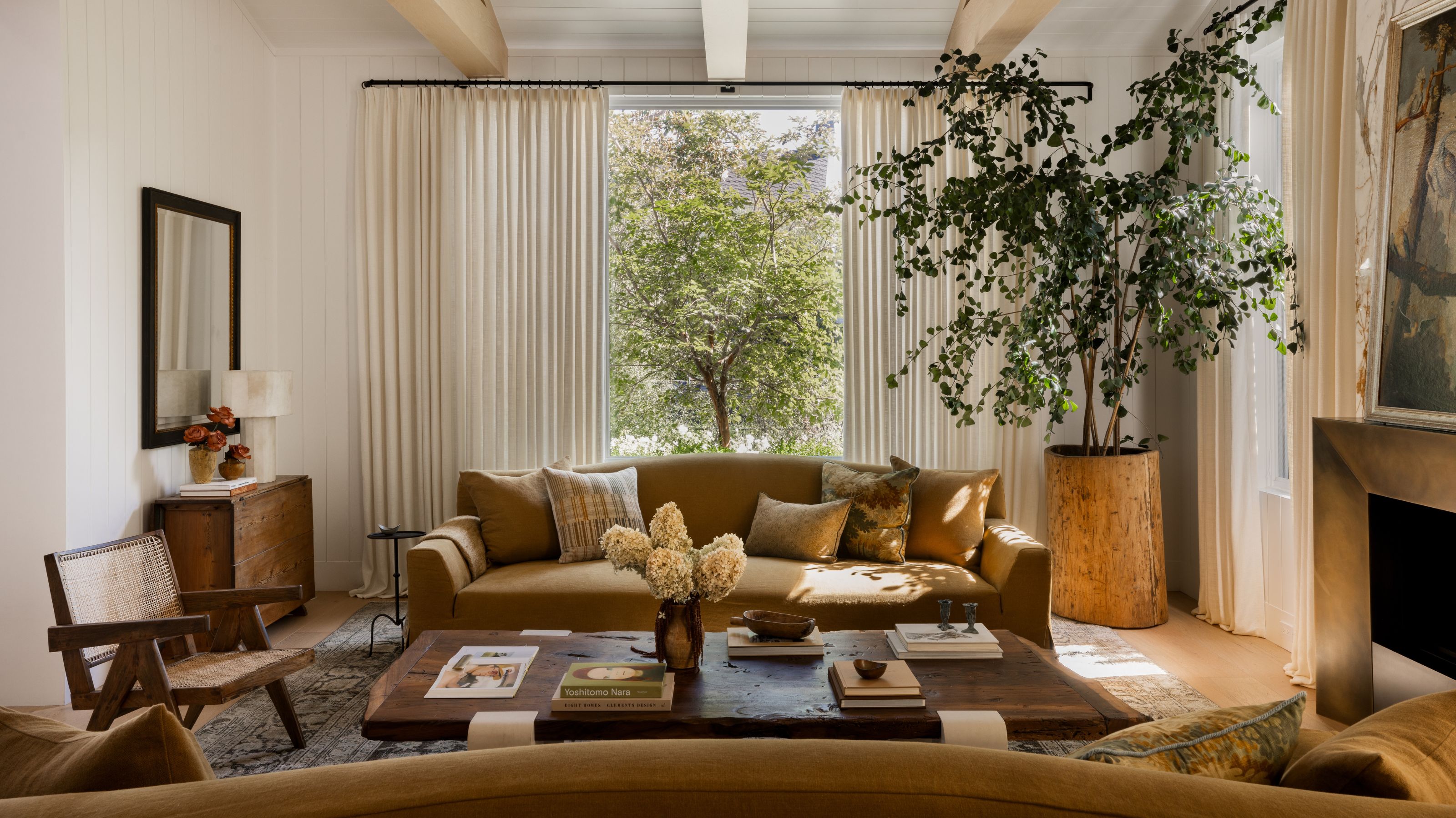 living room with wall of cream curtains, large potted tree, two ochre sofas opposite each other, a fireplace, timber coffee table styled with books, and a rattan armchair