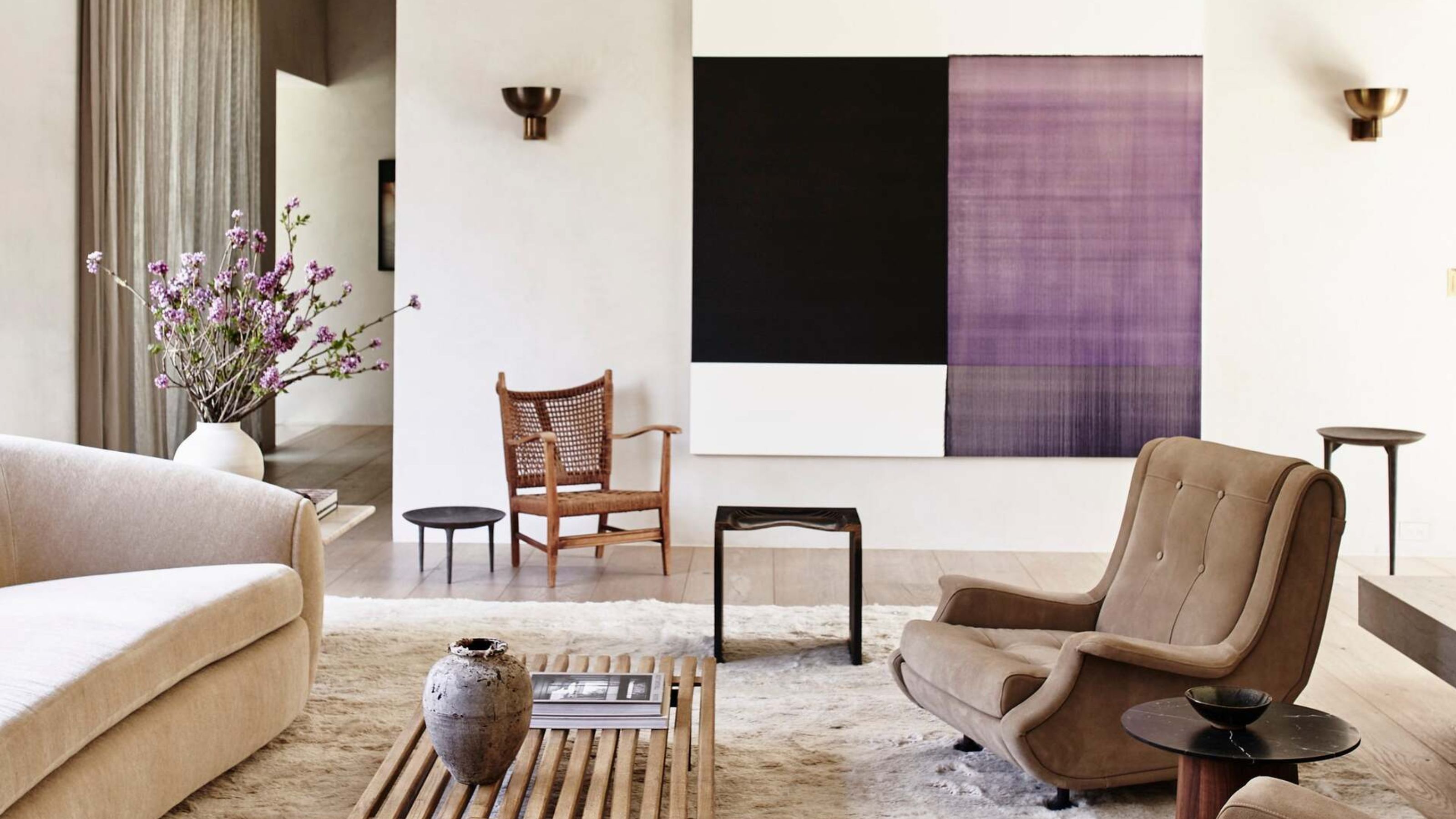 modern living room with white walls, sloped ceiling, modern chandelier, timber coffee table, white fluffy rug, brown armchairs, beige sofa, side tables, accent chair, and large artwork with sconces on each side