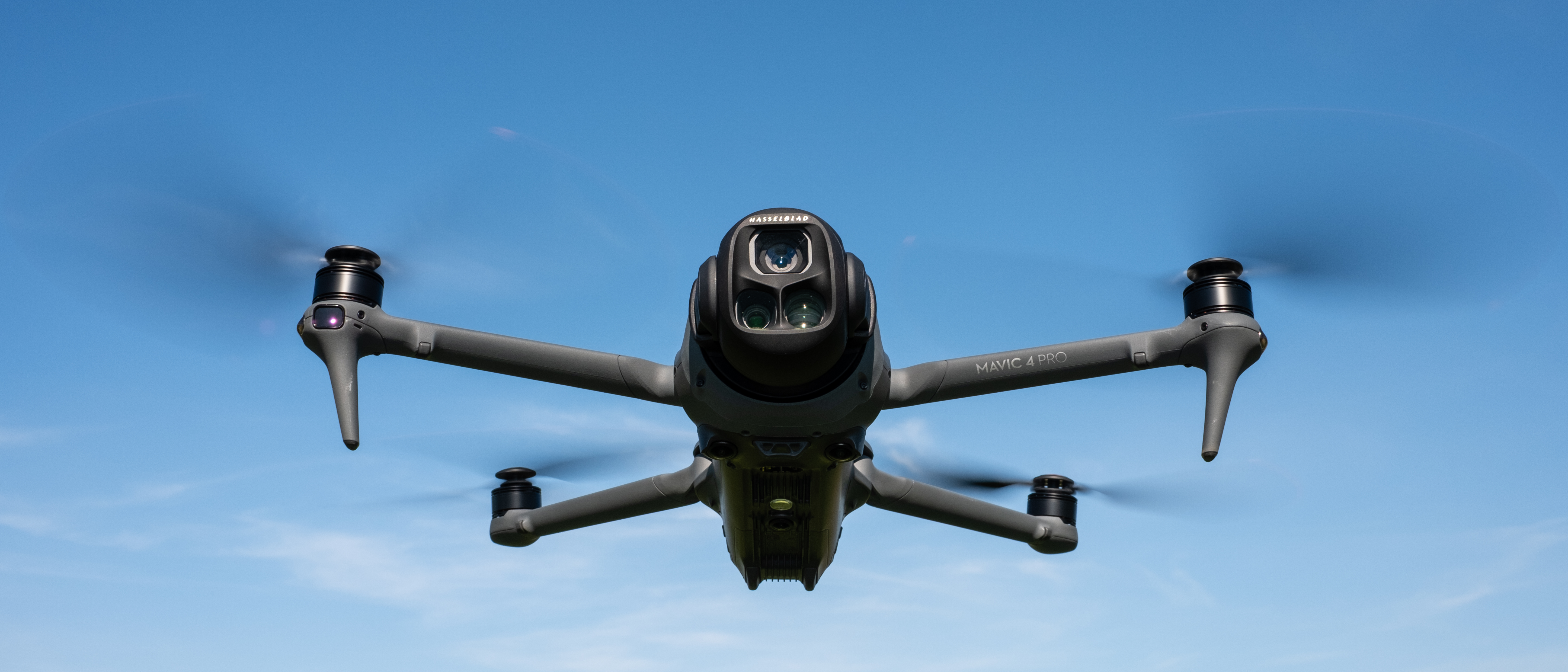 DJI Mavic 4 Pro in flight against a blue sky facing towards the camera with the drone camera and gimbal visible