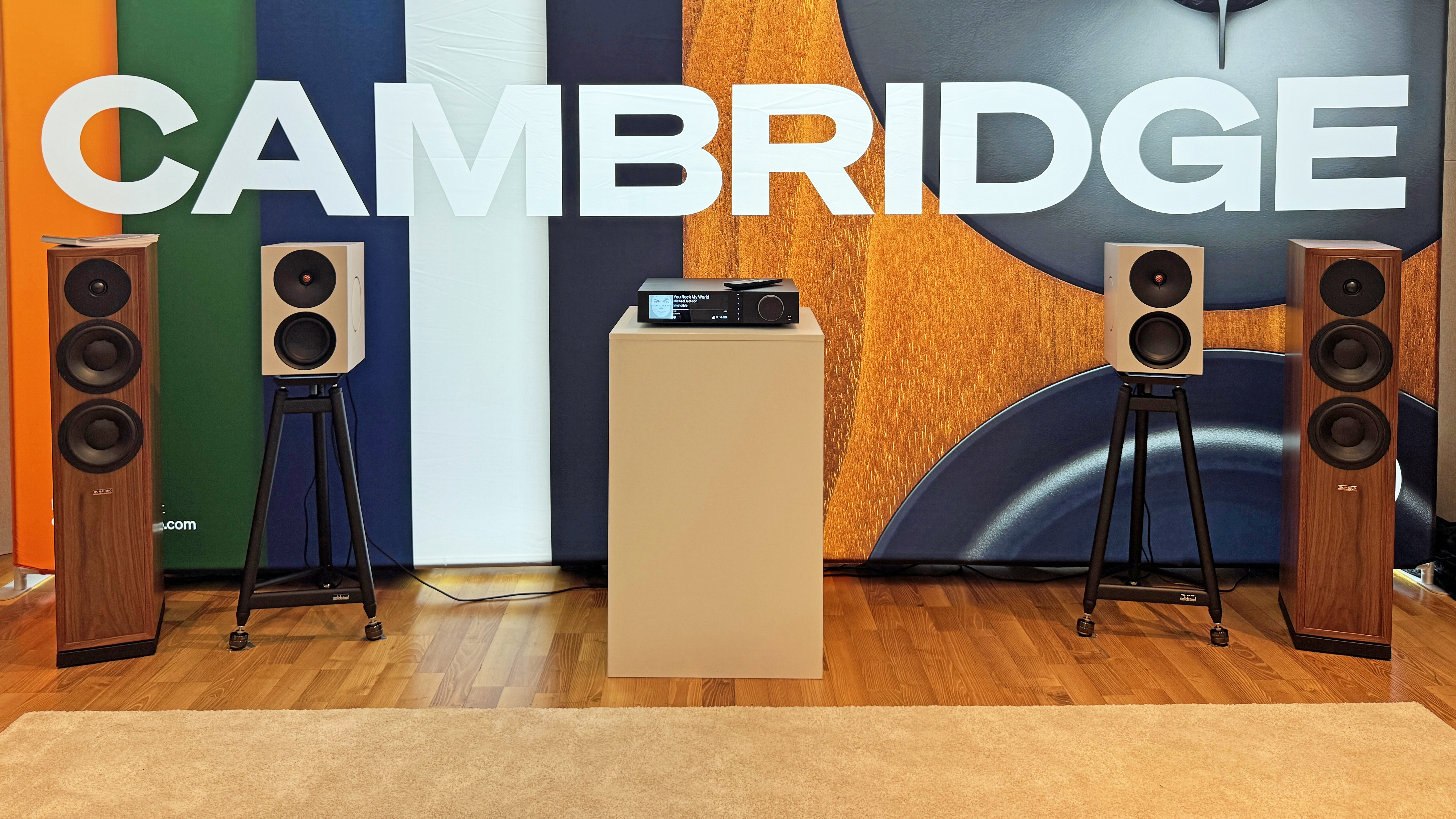 The Cambridge Evo 300 streamer at a trade show, sitting on a plinth between two large floorstanding speakers in a wood finish, plus a pair of smaller bookshelf speakers