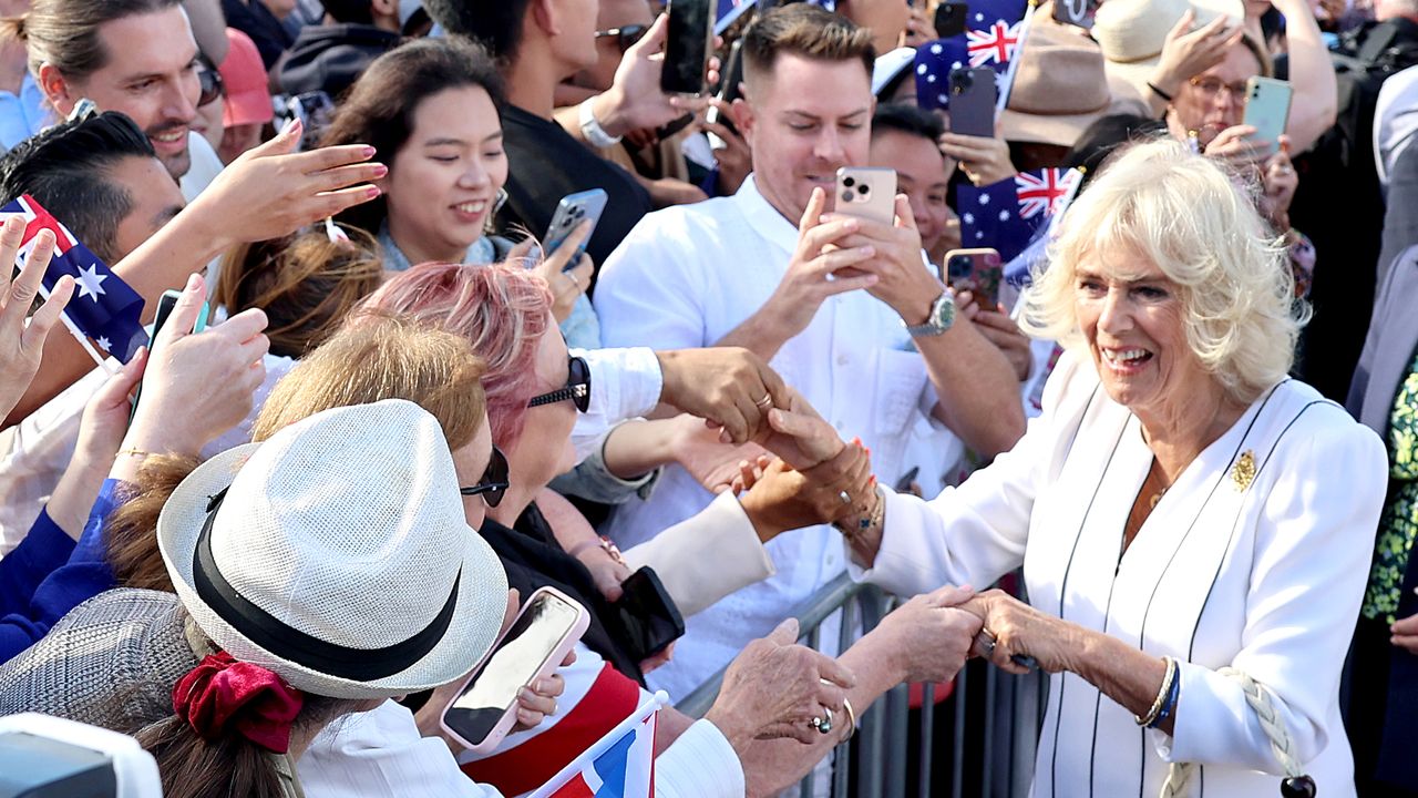 Queen Camilla wearing a white dress and shaking hands with a large crowd of fans waving Australian flags