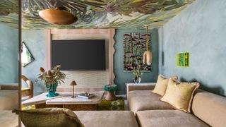 A living room interior with velvety beige seating topped with ochre cushions, a statement resin vase in bright blue and transparent acrylic, wavy wooden detailing, teal walls, and rattan decor.