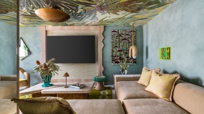 A living room interior with velvety beige seating topped with ochre cushions, a statement resin vase in bright blue and transparent acrylic, wavy wooden detailing, teal walls, and rattan decor.