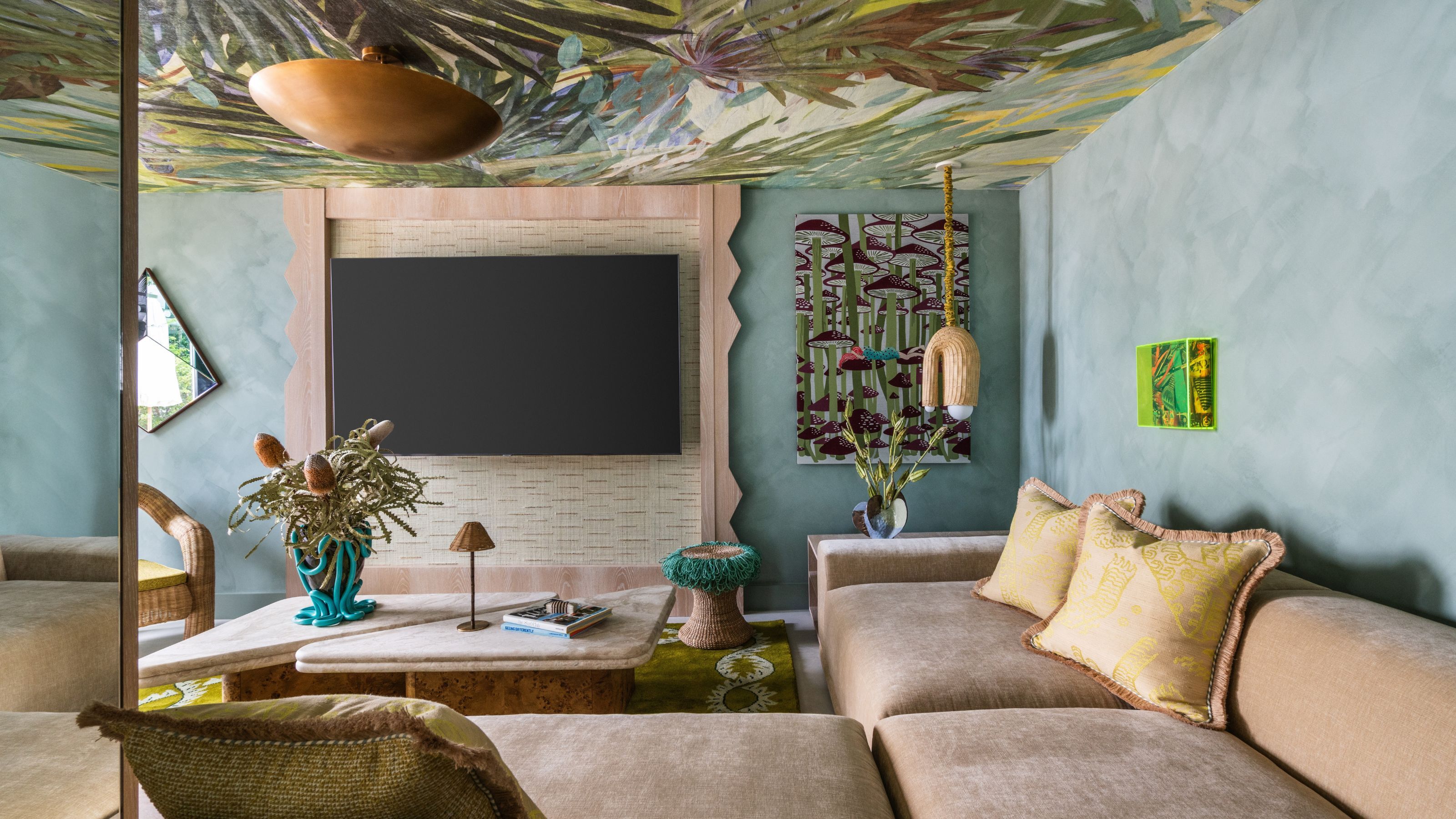 A living room interior with velvety beige seating topped with ochre cushions, a statement resin vase in bright blue and transparent acrylic, wavy wooden detailing, teal walls, and rattan decor.