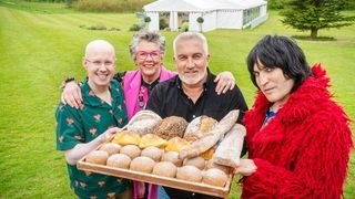 Matt Lucas, Prue Leith, Paul Hollywood, and Noel Fielding holding a tray of bread in front of the Bake Off tent