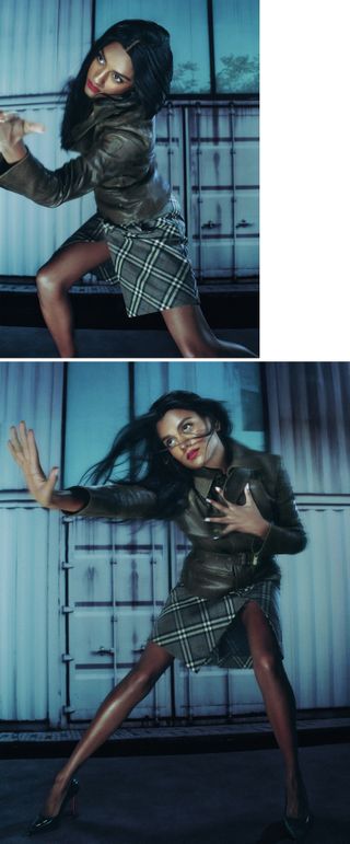 Simone Ashley is captured mid-movement in front of a metal crate backdrop wearing a green leather jacket and plaid pencil skirt.