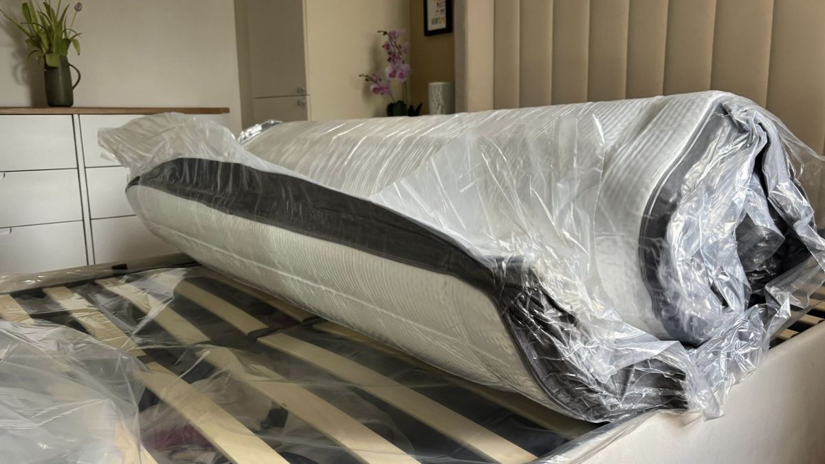 The image shows a dark grey and white mattress on a grey bed frame expanding inside its plastic wrapping