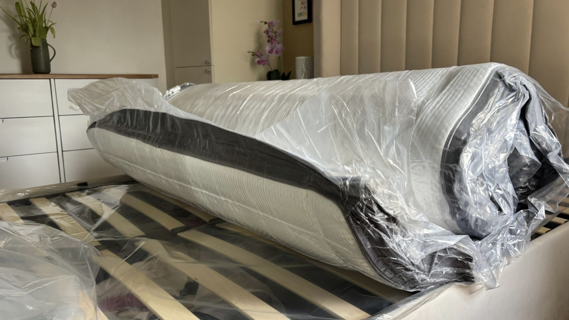 The image shows a dark grey and white mattress on a grey bed frame expanding inside its plastic wrapping