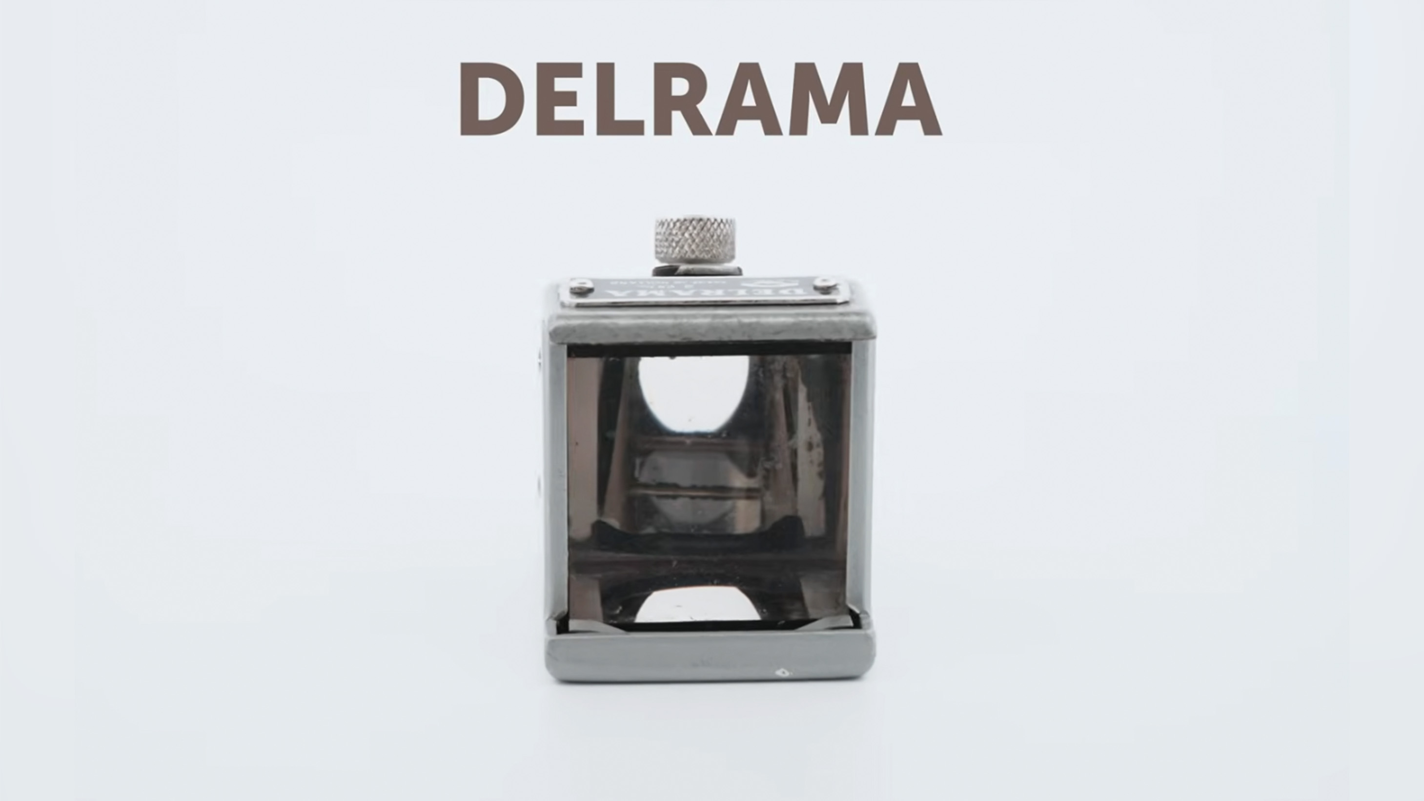 A small cube-shaped lens adapter for a camera. It has a glass front panel and a silver exterior. 