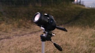 A Canon EOS Rebel T3i on a tripod aiming at the sky at night with grass and a fence in the background.