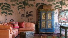 eclectic colorful living room with a folk style vintage cabinet, a large corner orange couch, and a pink mural wallpaper with tree and tropical print