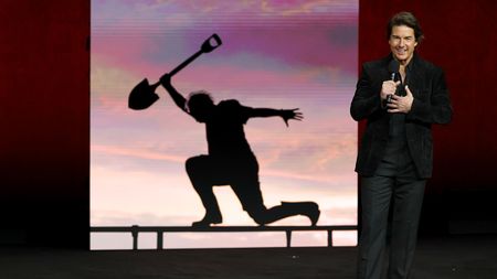 Tom Cruise in a black suit standing in front of a picture of his Digger character at CinemaCon 2026. 