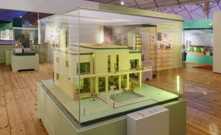View of a modern style doll house with a swimming pool, tennis court and multiple miniature people in a glass display case at the V&A Museum of Childhood. The space features wood flooring and green walls. Another doll house can be seen on display in the background