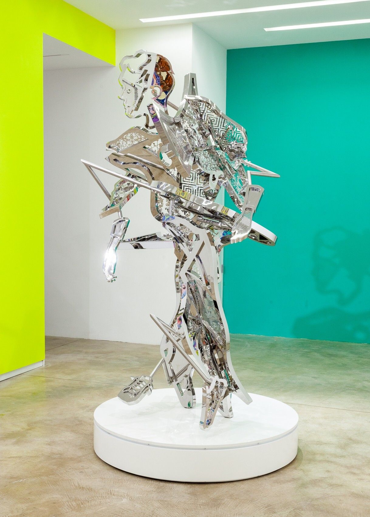A silver, anthropomorphic sculpture in shiny, different layers captures a man on the run while sitting on a white pedestal in a bright yellow and green room.