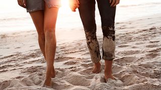 Couple holding hands and walking barefoot on the beach as the sun sets in the background.