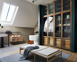 A dressing room with wooden and glass wardrobes next to an ensuite area with a sloped ceiling