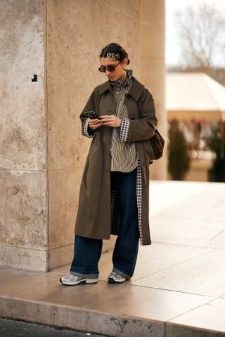 a guest at paris fashion week wears the sezane clyde trench coat with sneakers and wide leg jeans