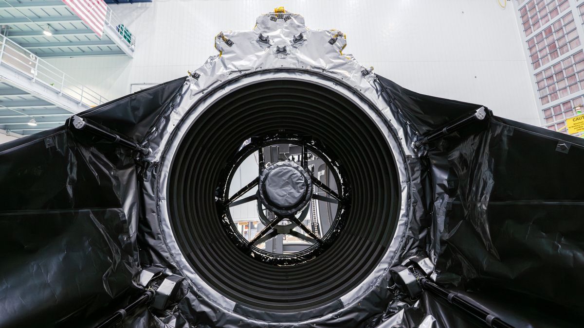 technicians in clean suits in a hangar next to a large rectangular spacecraft wrapped in metallic foil