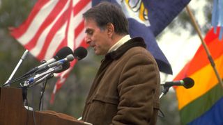 Carl Sagan speaks into a microphone at a podium with flags behind him