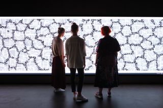 Marlies Miche, Elena Schwörer and Jeanette Ohlhäuser stand in front of a close up of the textile patterns