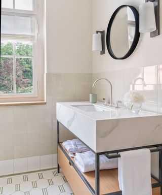 White marble bathroom vanity positioned near window with view, with small round black-framed mirror above