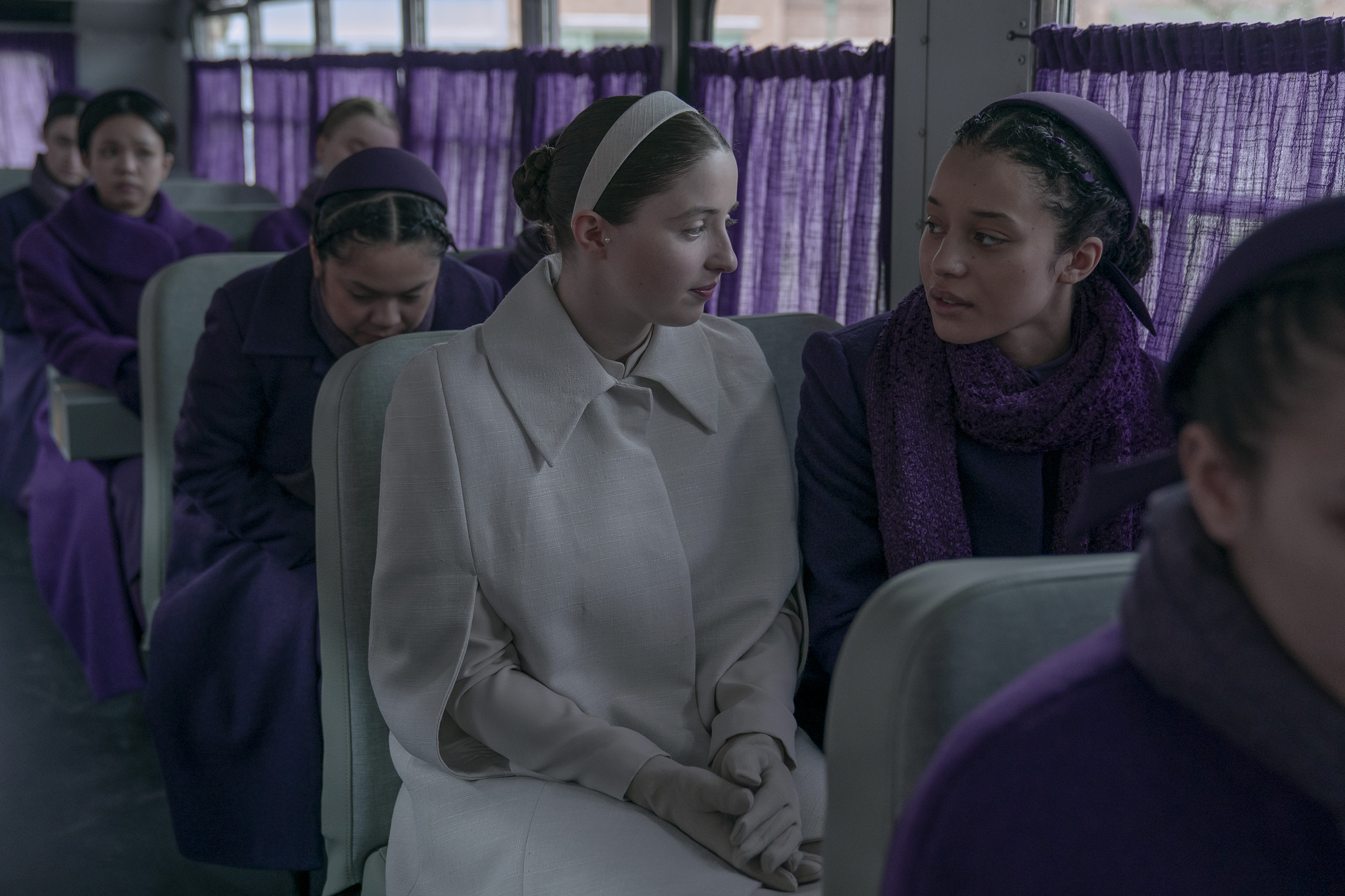 lucy halliday as daisy wearing her pearl girl white outfit and chase infiniti as agnes in a purple dress on a bus in the testaments