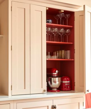 A peach kitchen cabinet opening up to three red shelves with glassware on them and a kitchen mixer at the bottom, on a white surface with peach lower cabinets underneath