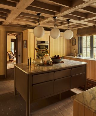 A log cabin kitchen with wooden ceiling beams, a stainless steel kitchen island toped with marble countertops, panelled wood cabinets and a tiled terracotta floor