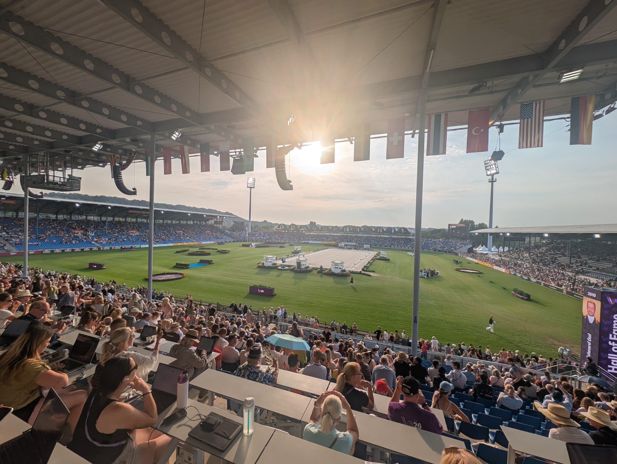 Image shows Aachen's main arena in the evening light &amp;ndash; in the distance is a sand dressage arena surrounded by grass, in the foreground are full stands