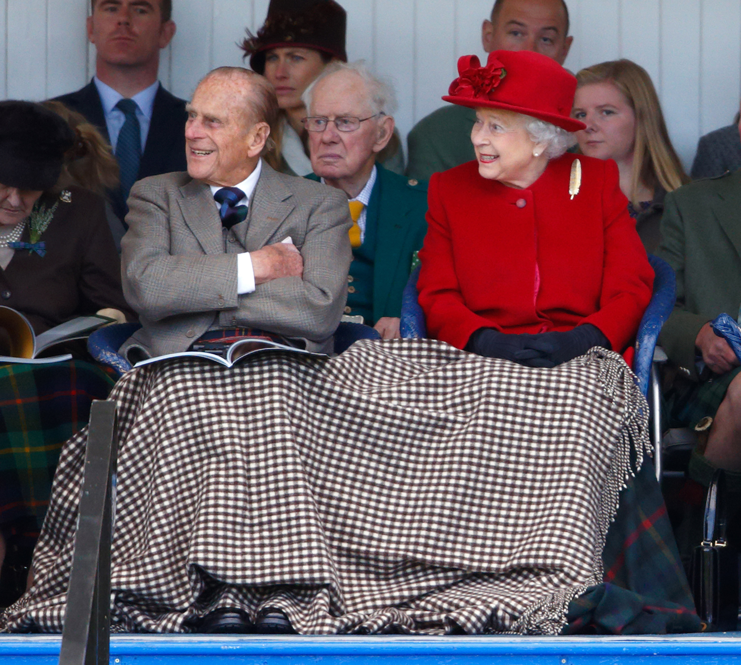 Prince Philip and Queen Elizabeth, wearing a red coat and hat, sitting under a blanket smiling in Scotland