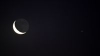An upturned crescent moon glows to the left of the star-like light of Mars in a black sky.