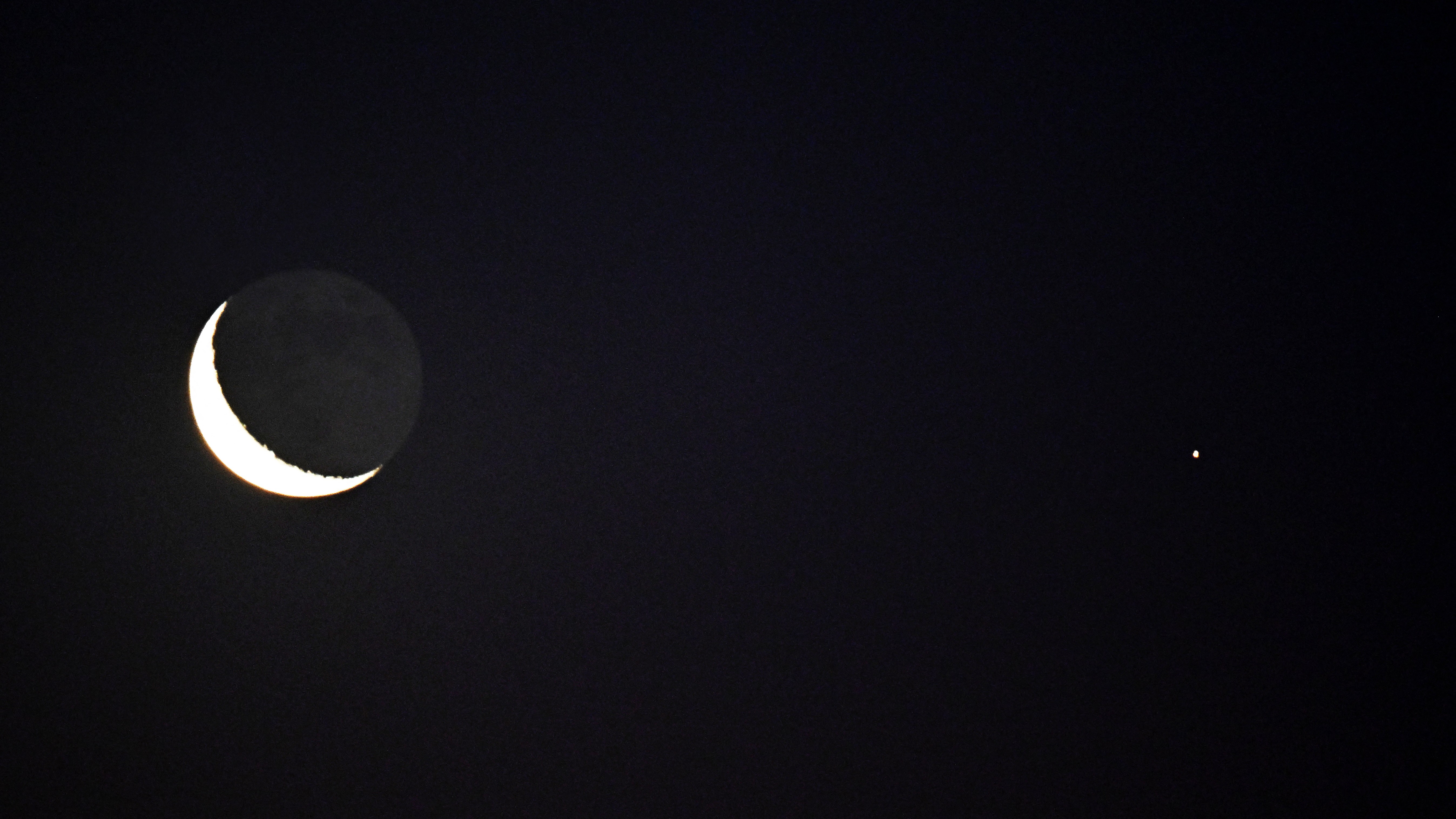 An upturned crescent moon glows to the left of the star-like light of Mars in a black sky.