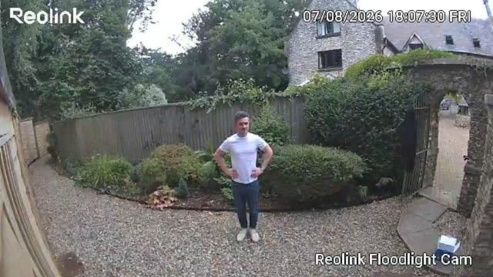 Reolink camera footage of man standing on gravel pathway beside fence, garden and house in background.