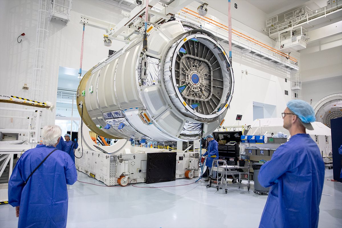 a cylindrical silver spacecraft inside a white-walled high bay