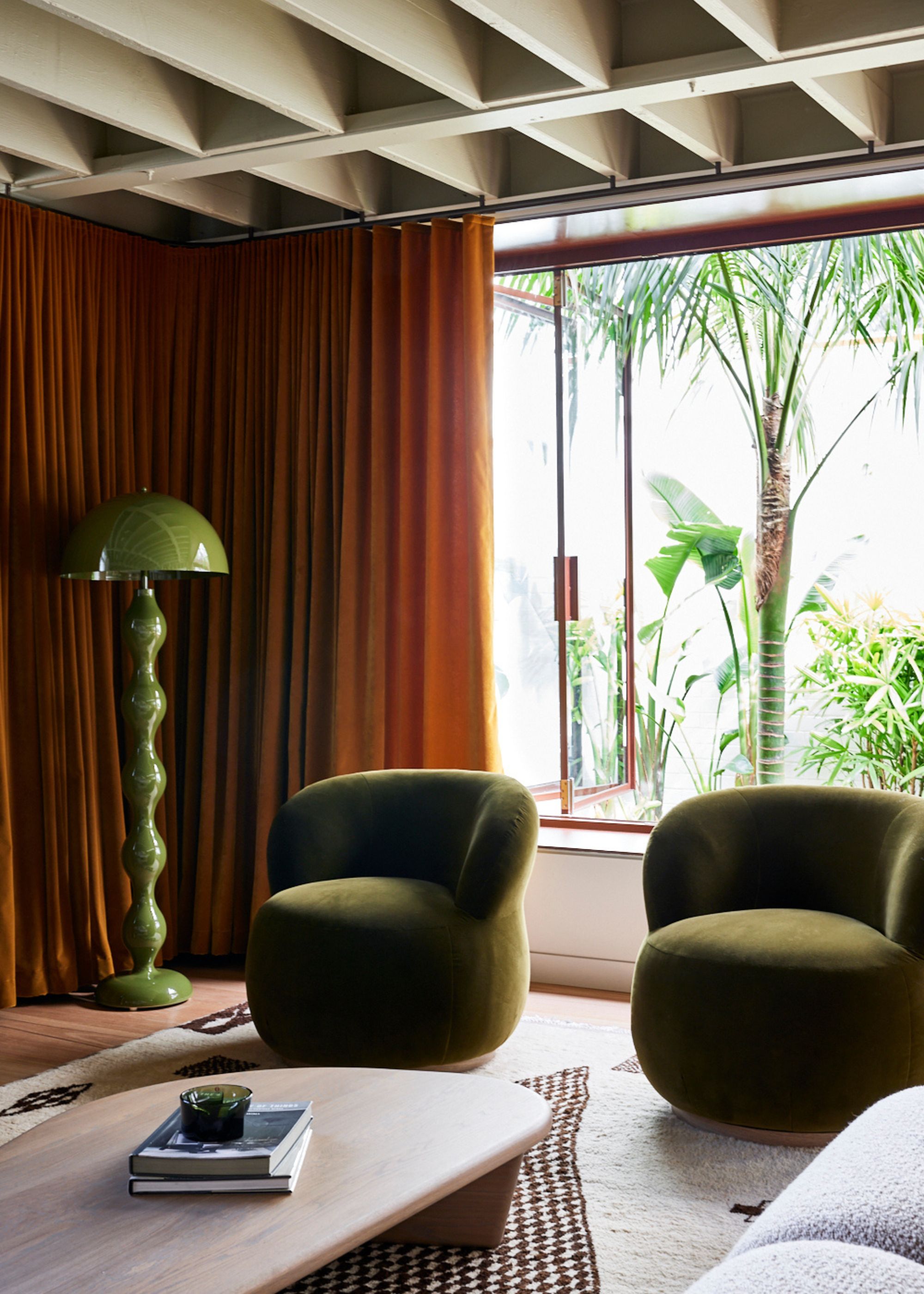 A living room with two green accent chairs beside a window as well as a large coffee table in the middle of the room. There are also floor to ceiling windows with large curtains draping down. Next to the curtain is a tall green floor lamp.