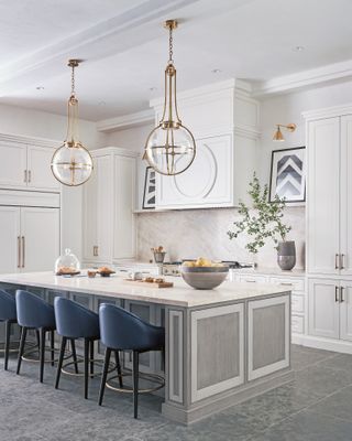 A luxe white kitchen with gray island, blue chairs, gold accents in the door handles and statement pendant lighting.