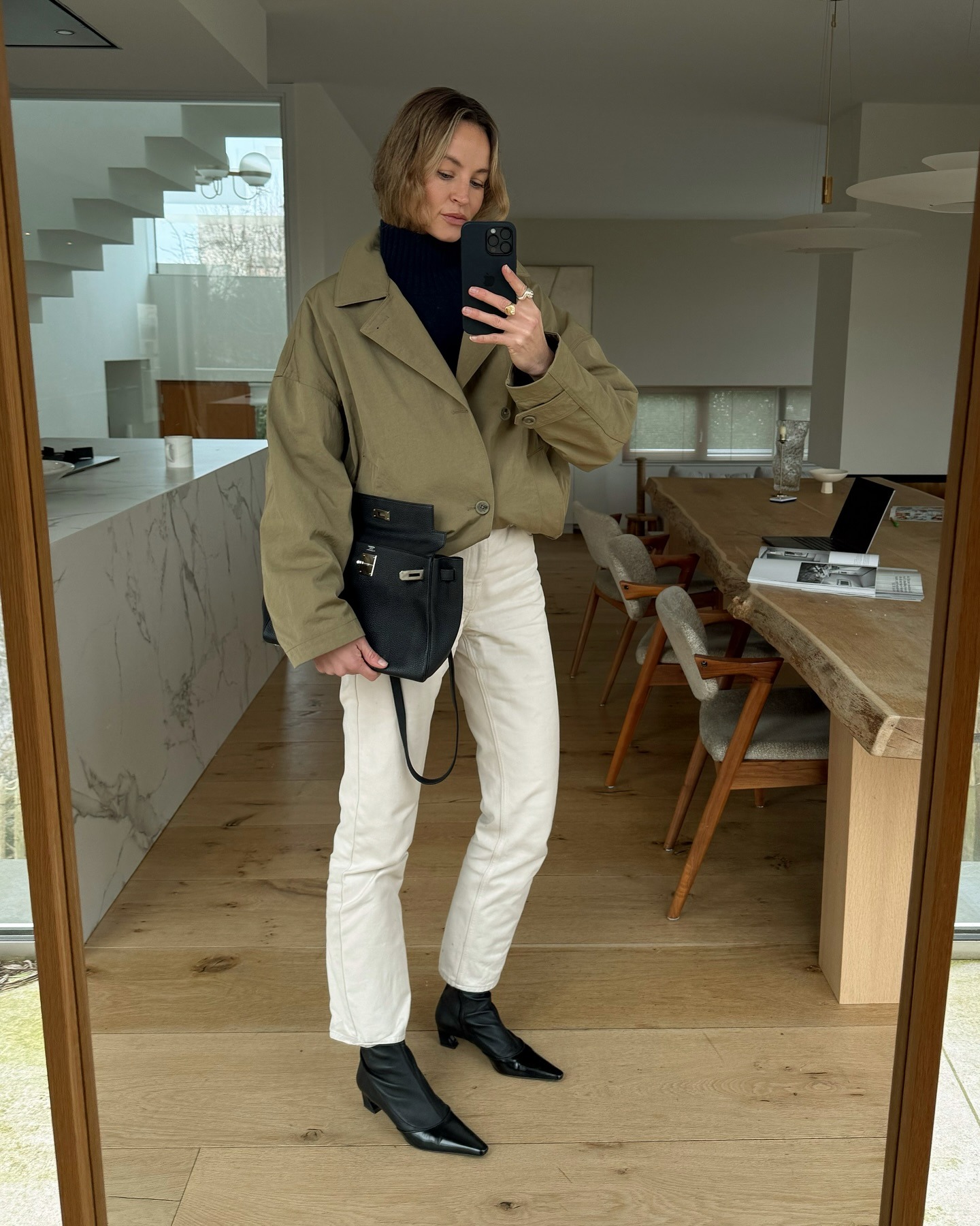 Anouk wears a black turtleneck, biege coat, white jeans, and black boots.
