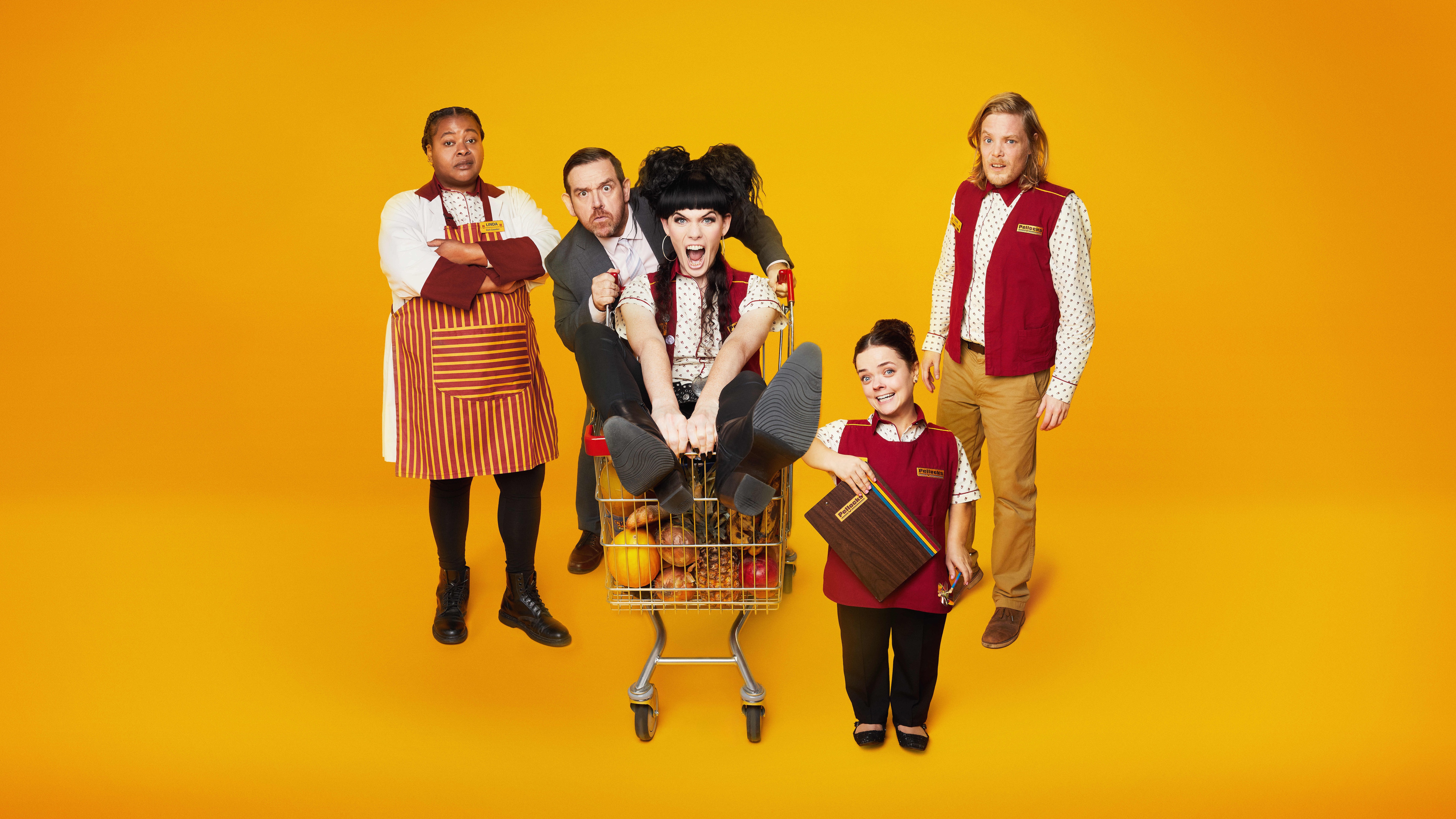 A key art image for the ITV sitcom Transaction. The image shows Liv (Jordan Gray) riding on top of a supermarket trolley with her feet pointing towards the camera, while Simon (Nick Frost) is holding on to the trolley&#039;s handle behind her. On the left stands Beefy Linda (Kayla Meikle), wearing a supermarket uniform with a red and yellow striped apron over the top, standing with her arms folded and looking bored. On the right in the foreground stands Millie (Francesca Mills), also in a supermarket uniform, smiling brightly and holding a clipboard, and behind her is Tom (Thomas Gray), in a supermarket uniform with a neutral expression.