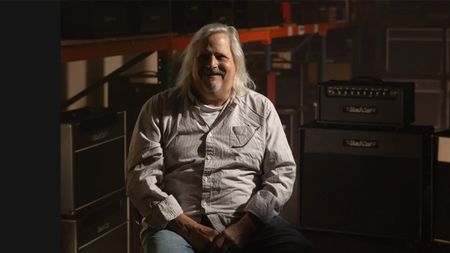 Mark Sampson sitting on a stool and smiling at the camera, with amps in the background