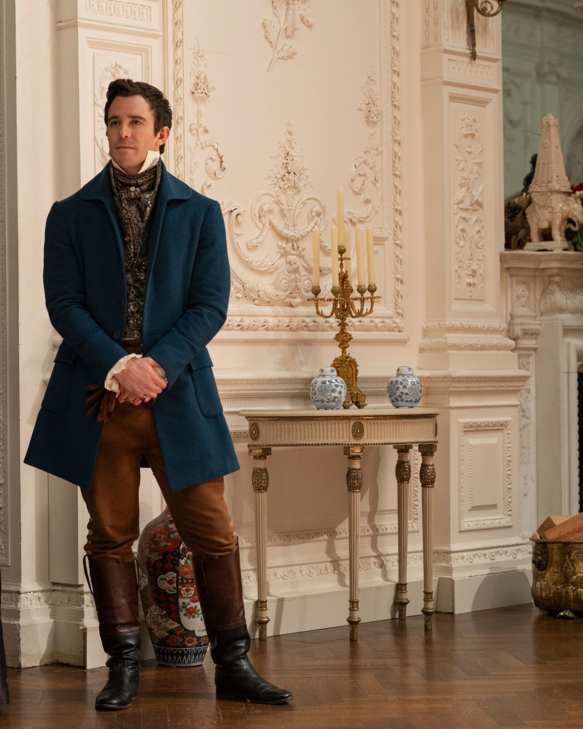 Production still from Netflix's Bridgerton Series showing a man in a blue jacket and brown pants and boots standing in front of a white wall that has elaborate crown molding on it.