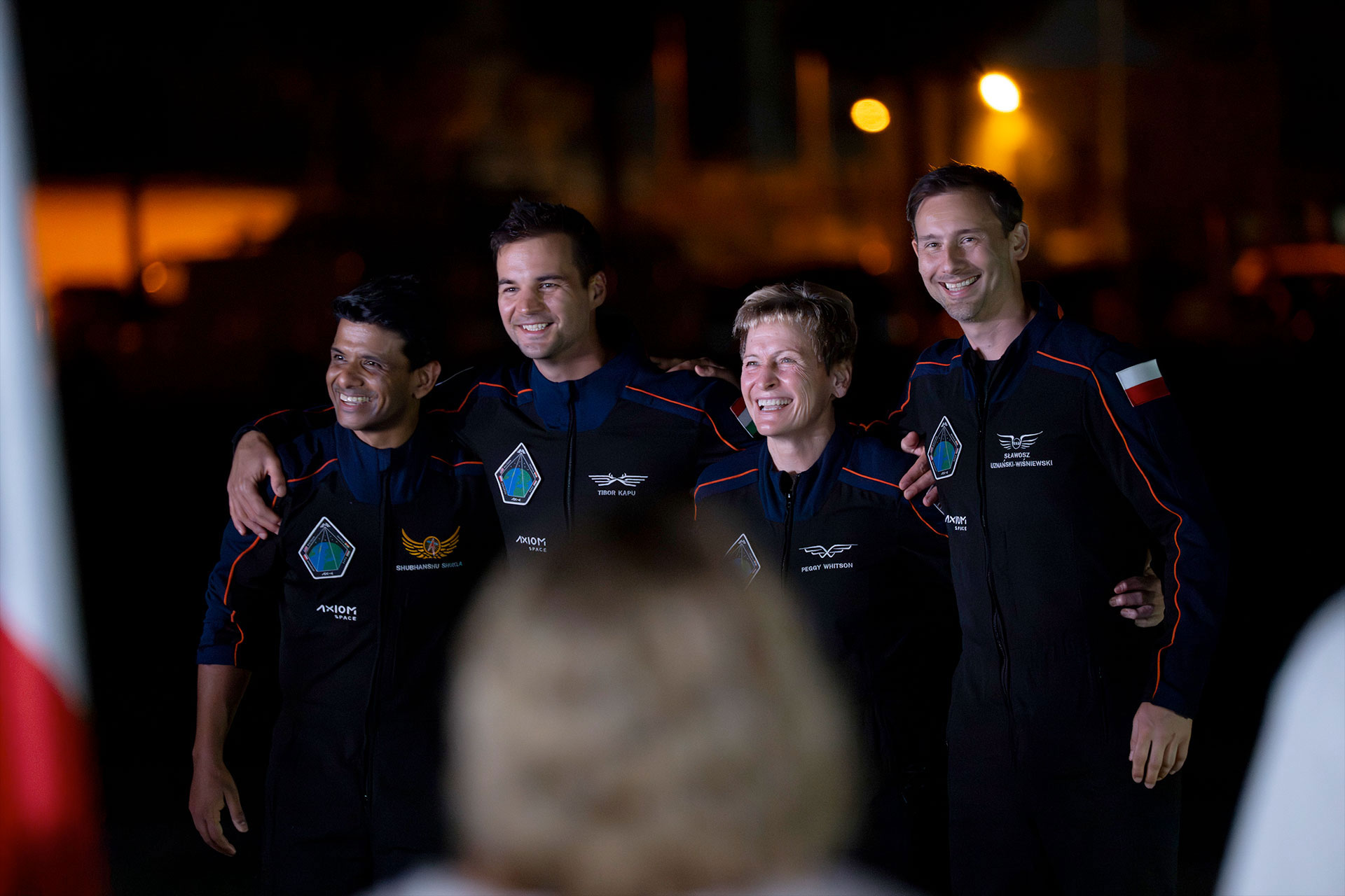 four people wearing dark blue flight suits pose together outside at night for a photo