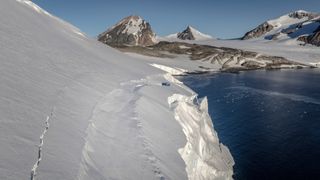 a snow-covered glacier meets the sea, with rocky mountains visible in the background