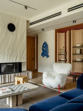 modern living room with textured fireplace, coffee table with chess board, gray rug, blue sofa, and fluffy white armchair with red legs