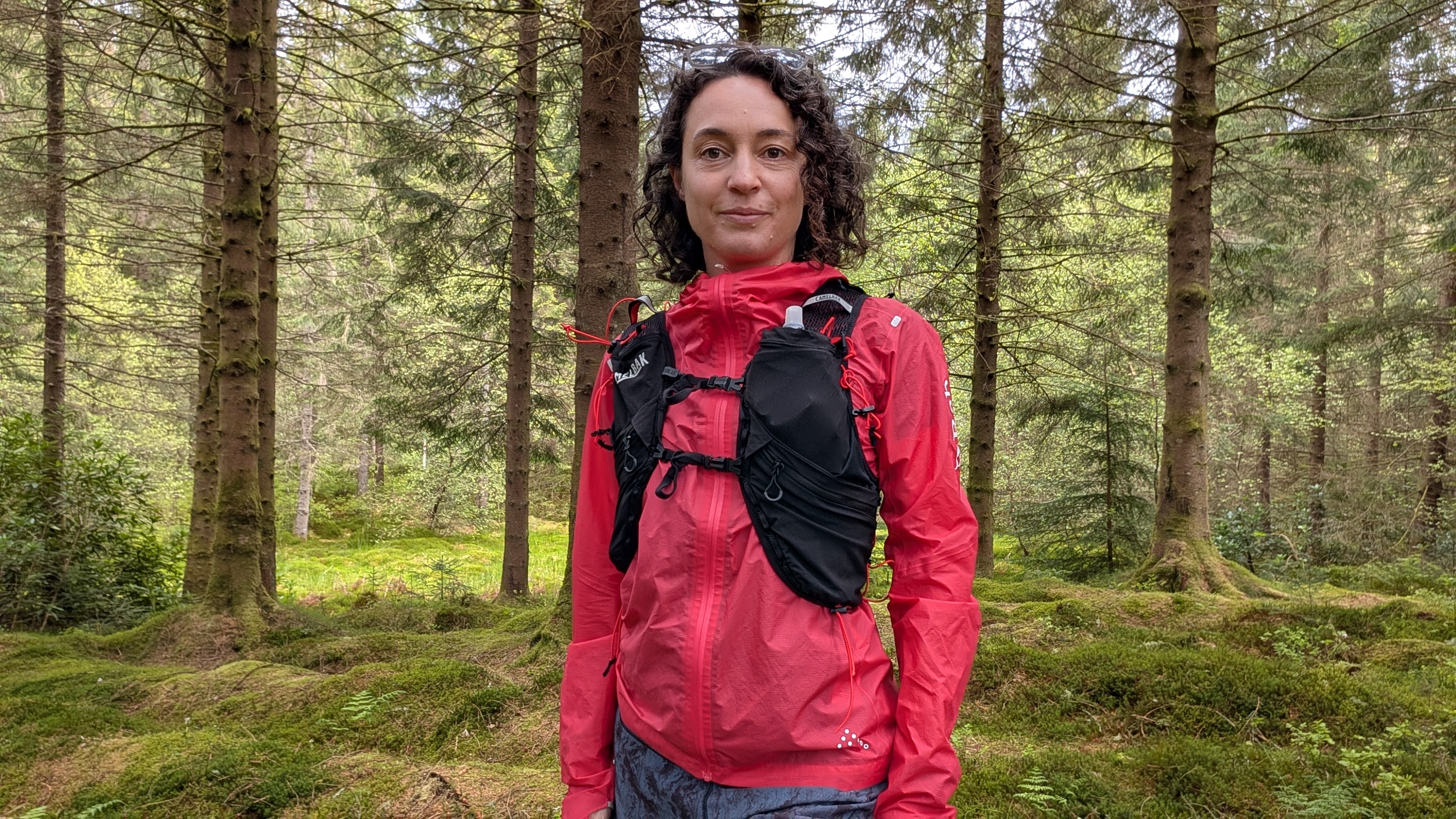 A woman trail runner wearing a pink jacket and black Camelbak Apex Pro running vest in the forest