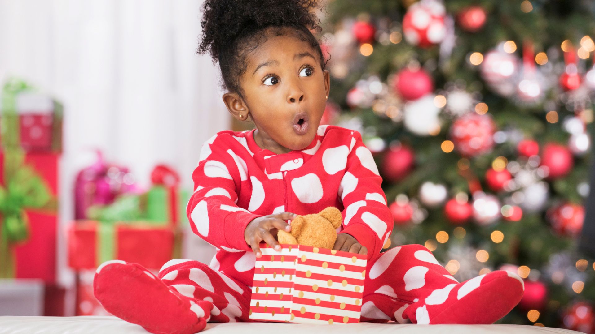 A little girl unwraps a gift in front of a Christmas tree with a look of surprise on her face