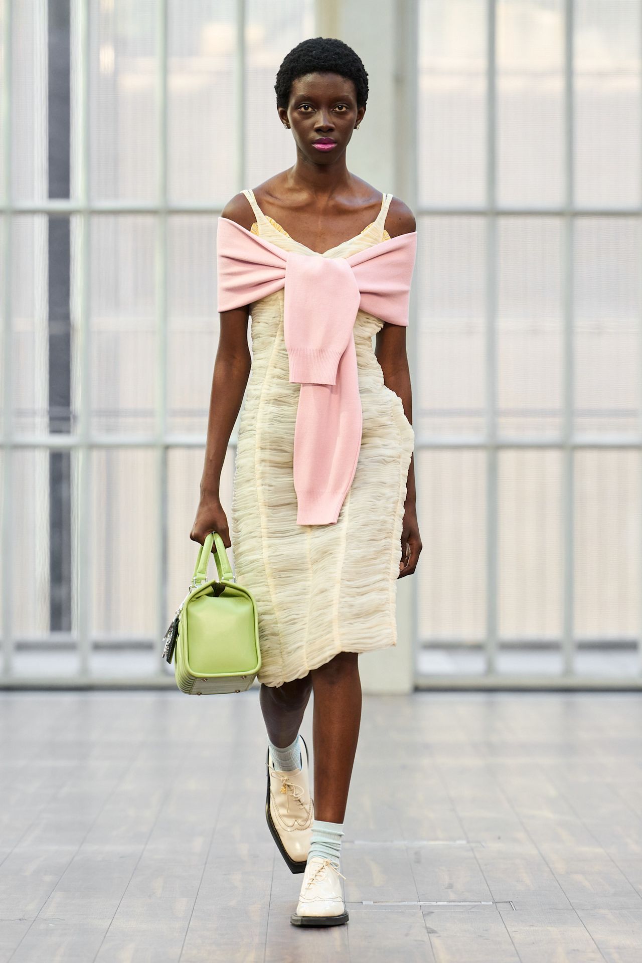Layering staples for spring 2026 is shown in a photo from Mithridate's runway show, with a model walking while wearing a pale yellow dress styled with a pink sweater tied around the shoulders, a bright green bowler bag, light blue socks, and pale yellow derby shoes