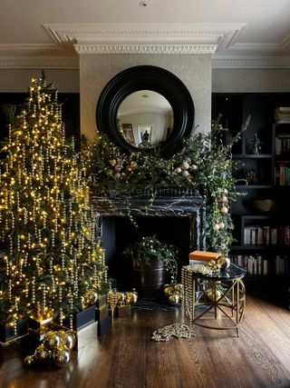 Dark living room with Christmas tree covered in gold beaded decorations and lights, with dramatic green foliage garland over mantlepiece
