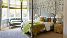 Four-poster bed in opulent room with patterned green wallpaper. The bed has neatly tucked in sheets, lots of throw pillows and a chesterfield sofa in the bay window 